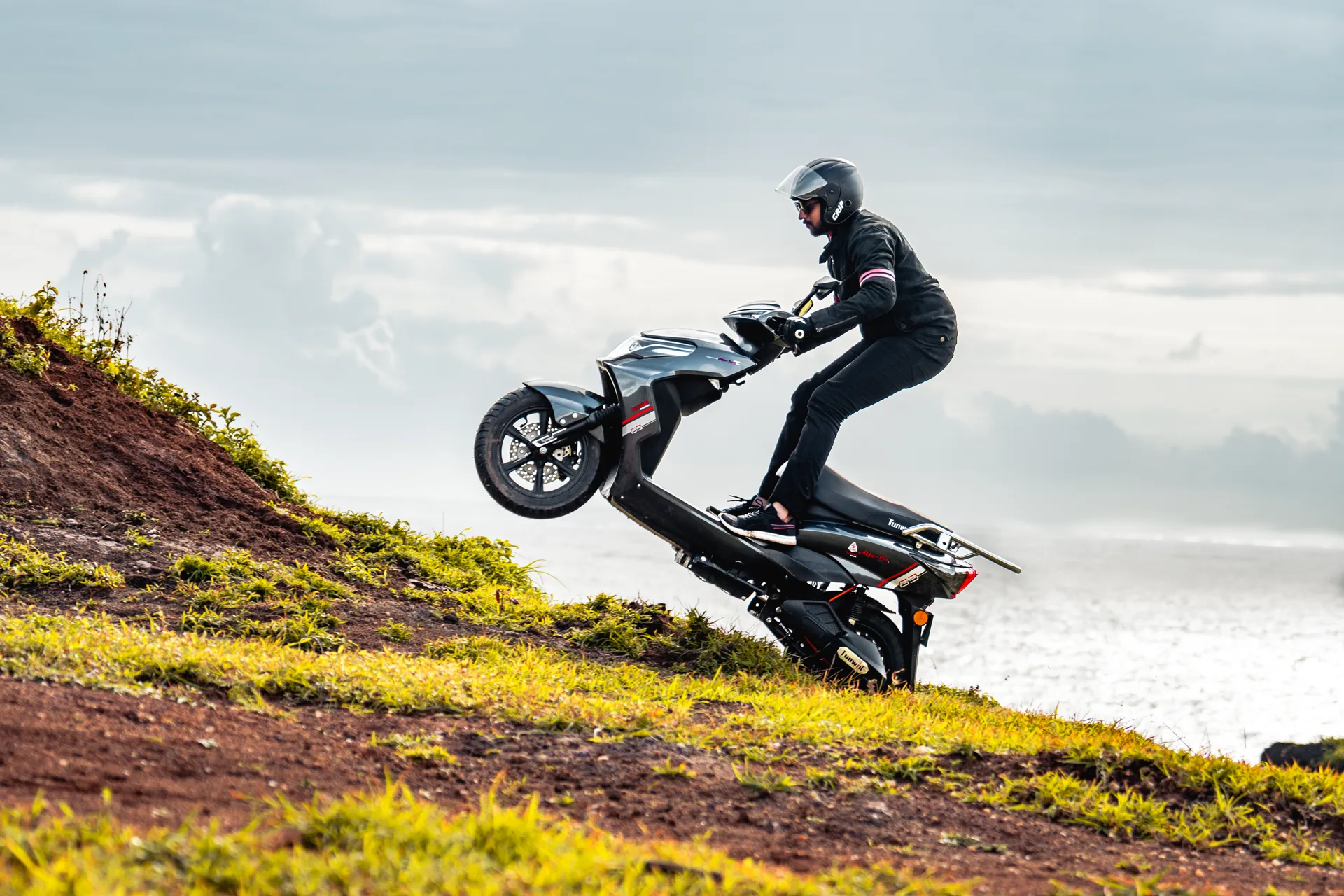 A rider lofting the front wheel of a Tunwal scooter on a coastal hill